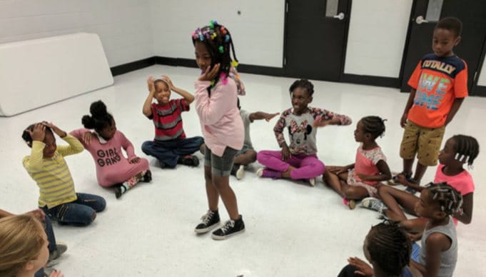 elementary-aged children sit in a circle on the ground during a drama game